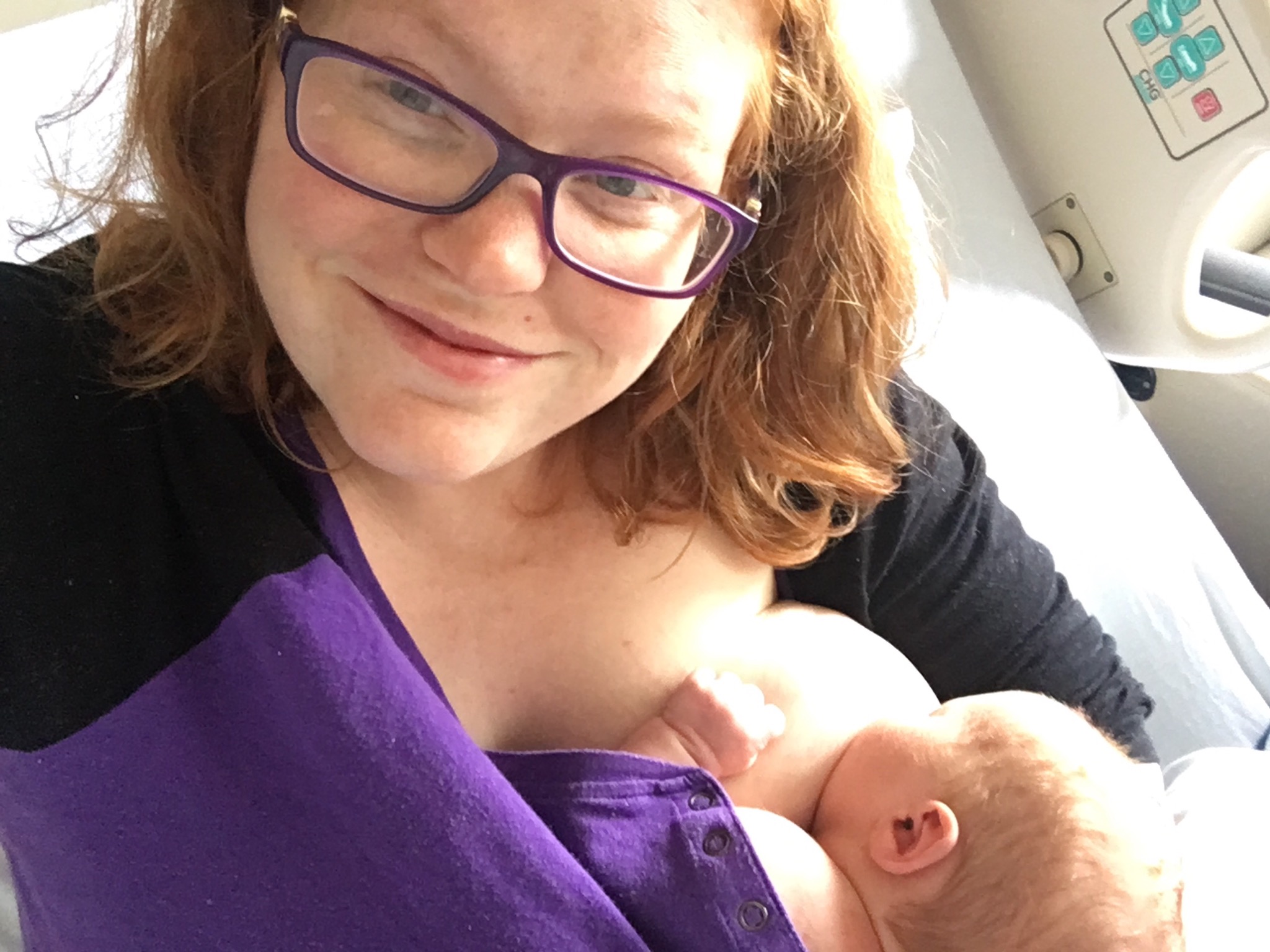 mother with red hair and wearing glasses smiles at camera as preemie infant breastfeeds