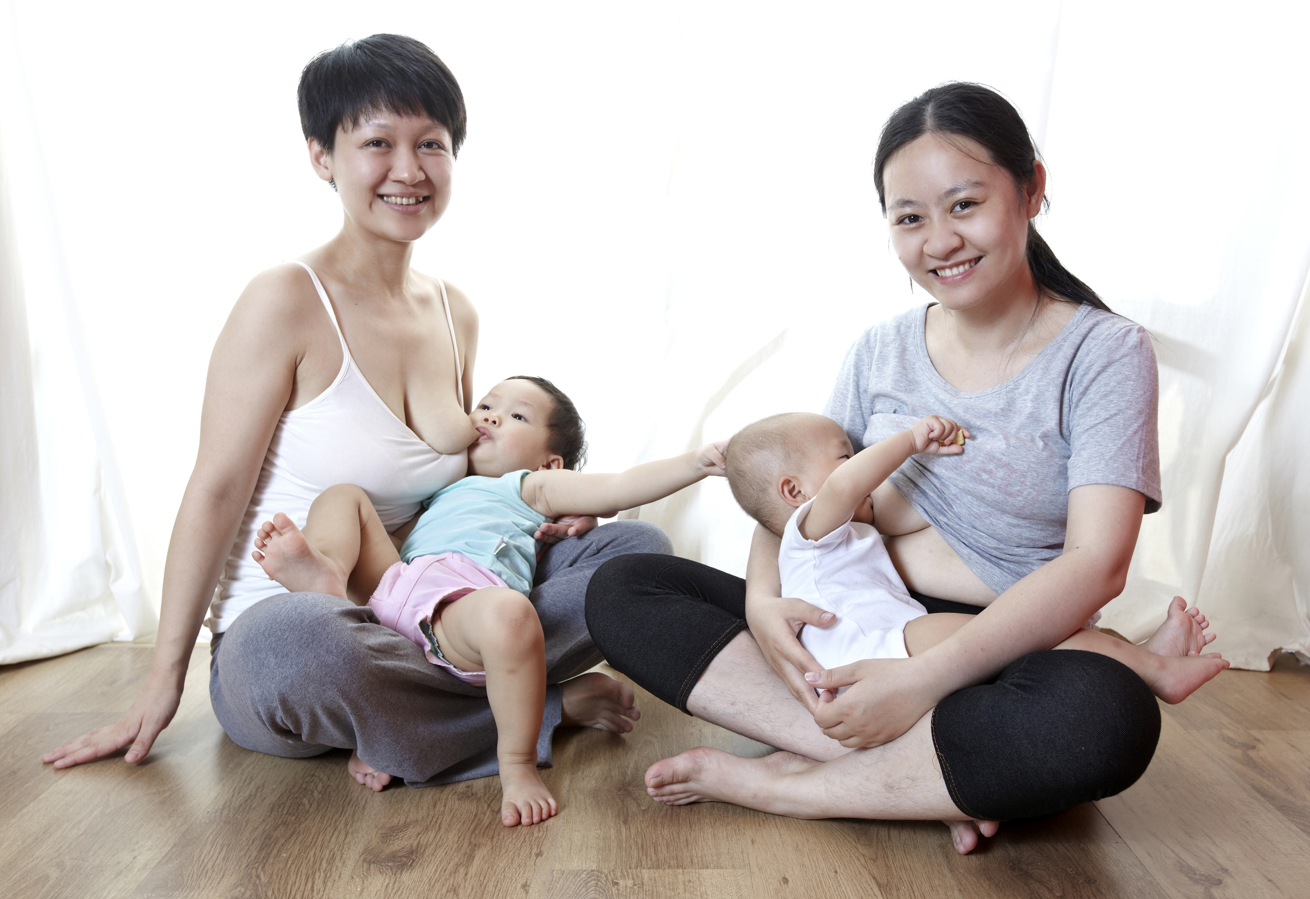 two mothers smiling while seated on a floor and breastfeeding their infants. one infant reaches for the other